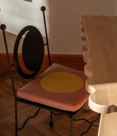Metal chair with a black circular backrest and a pink seat cushion featuring a large yellow circle, next to a wooden table with a scalloped edge.