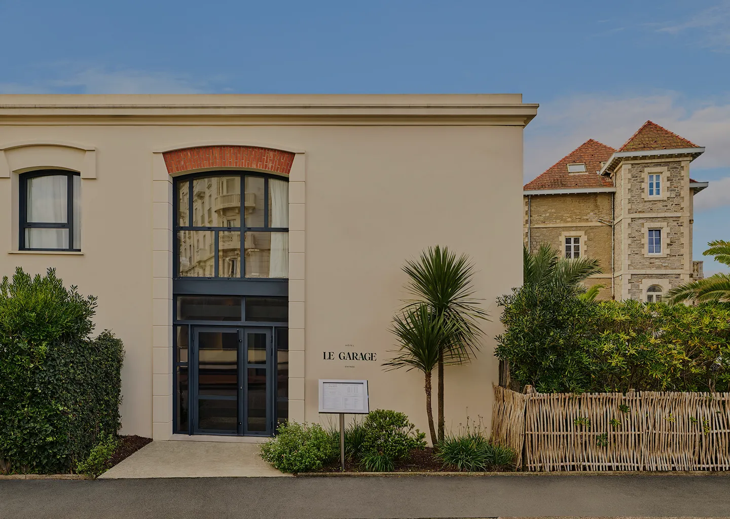 Entrance to Hôtel Le Garage with large window and door framed by beige walls, greenery, and a signboard in front.