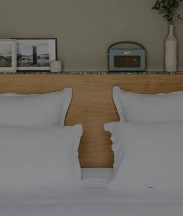 Two white pillows on a bed with a wooden headboard, a vintage-style radio, framed photos, and a vase on a shelf above.