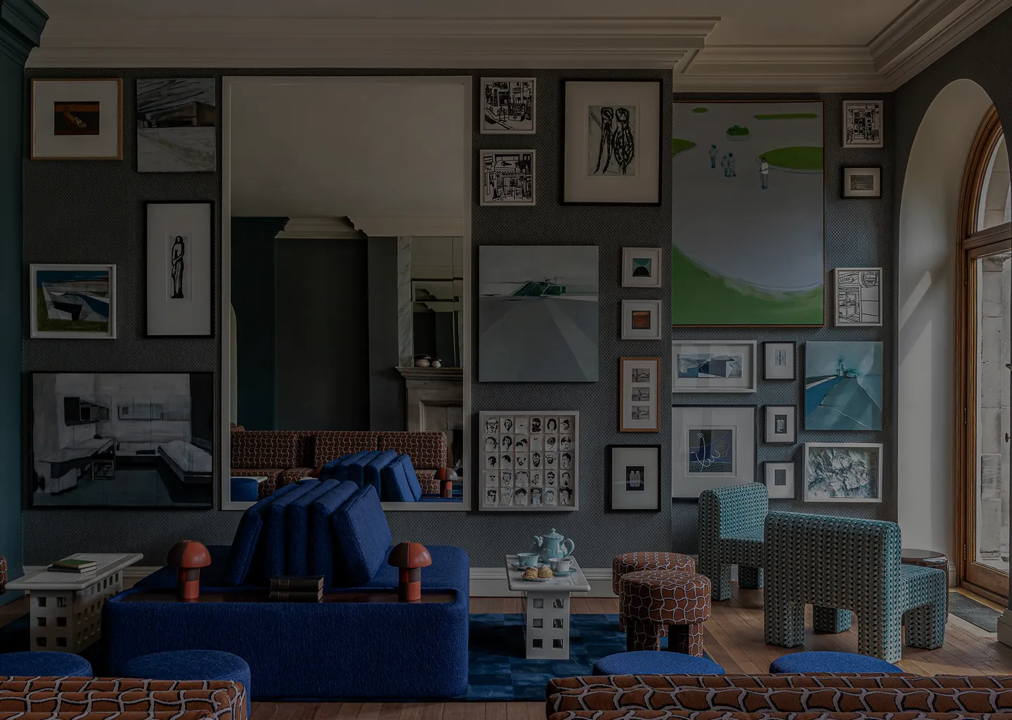 Modern living room with blue and patterned furniture, a gallery wall of framed art, and a large window with an arched wooden frame.