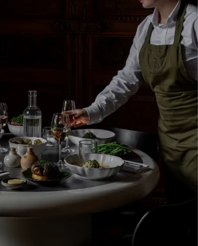 Person in an olive green apron serving wine at a table set with plates of food and a glass water bottle.