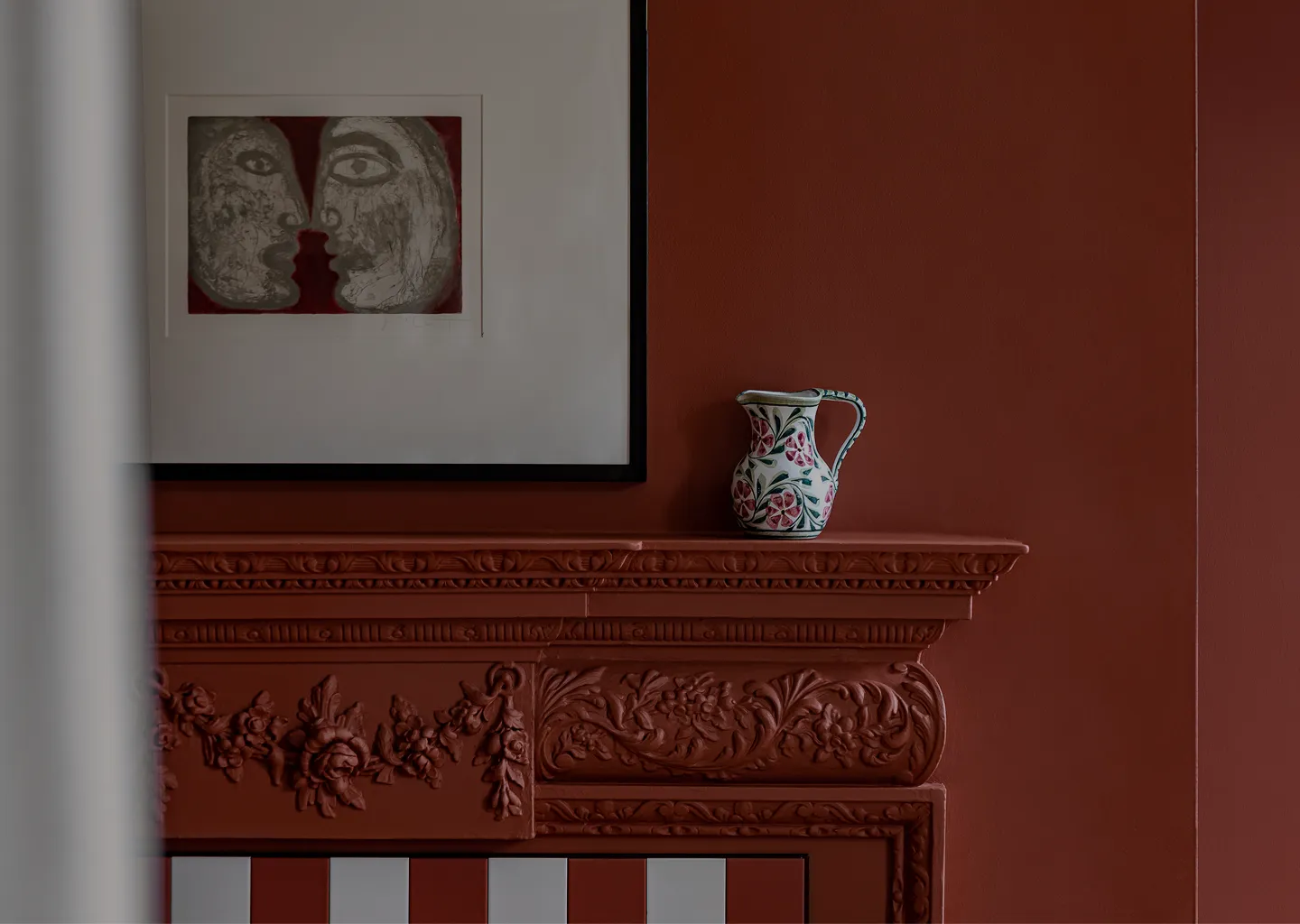 Decorative ceramic pitcher with floral patterns on an ornately carved reddish-brown mantel beneath a framed abstract art print of two faces.