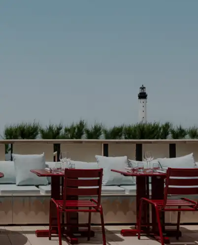 Outdoor seating area with red chairs and tables, white cushions on a bench, and a lighthouse visible beyond greenery under a clear sky.