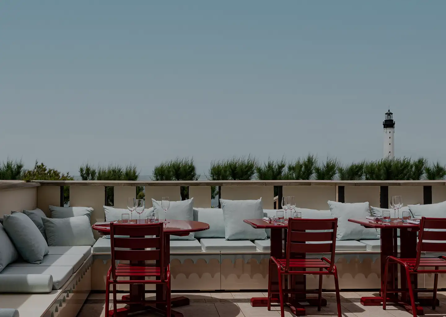 Outdoor seating area with red tables and chairs, gray cushioned benches, and a lighthouse visible in the distance under a clear sky.