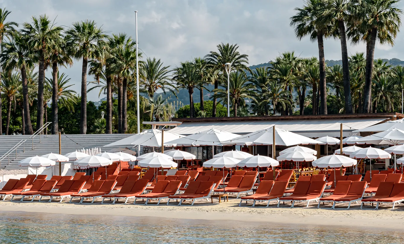 Rows of empty orange beach lounge chairs with white umbrellas on a sandy beach with palm trees and a building in the background.