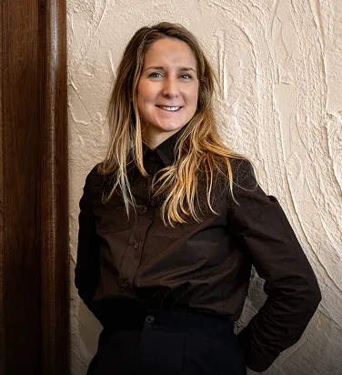 Smiling woman with long blonde hair wearing a black shirt, standing against a textured white wall.