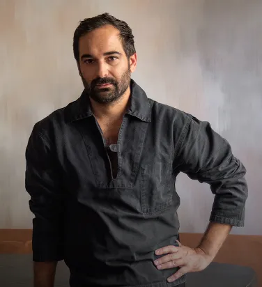 Bearded man wearing a dark long-sleeve shirt with one hand on his hip against a neutral background.