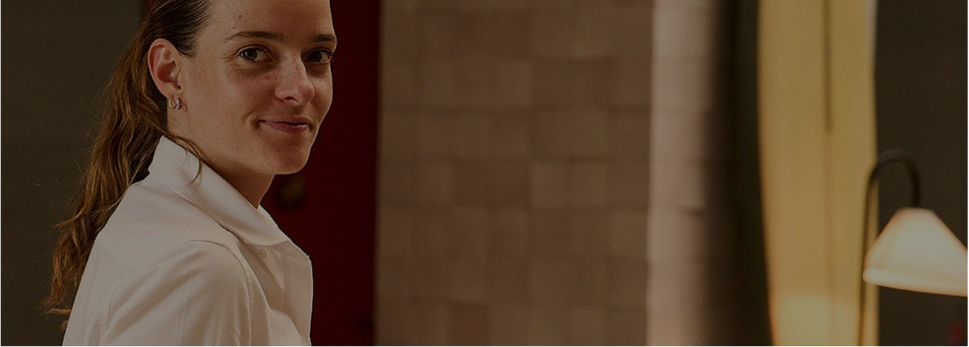 Woman wearing a white shirt smiling slightly while looking toward the camera in a softly lit room.