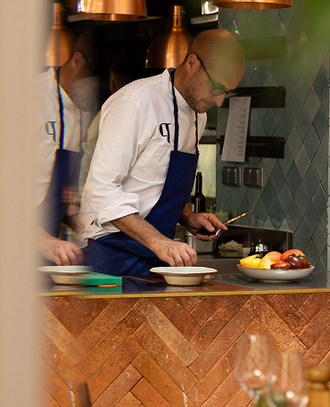 Chef in a white jacket and blue apron carefully preparing dishes in a kitchen with tiled walls and a wooden counter.