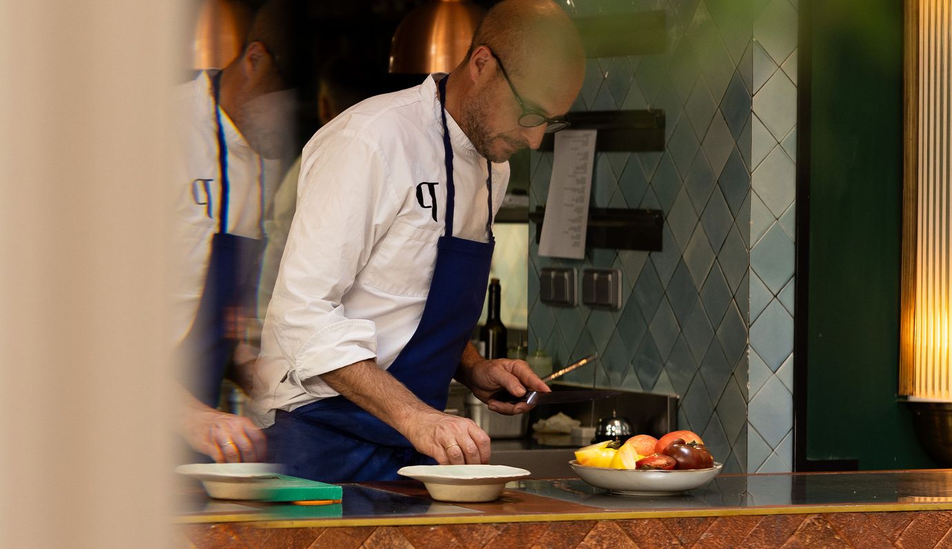 Chef wearing glasses and a blue apron preparing food in a kitchen with a bowl of vegetables on the counter.