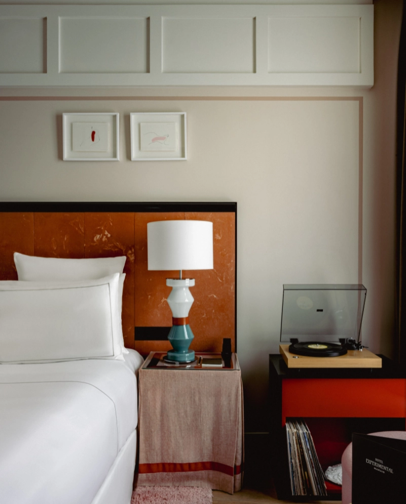 Modern bedroom corner with white bed, decorative lamp on a side table, and a turntable on a red-black cabinet.