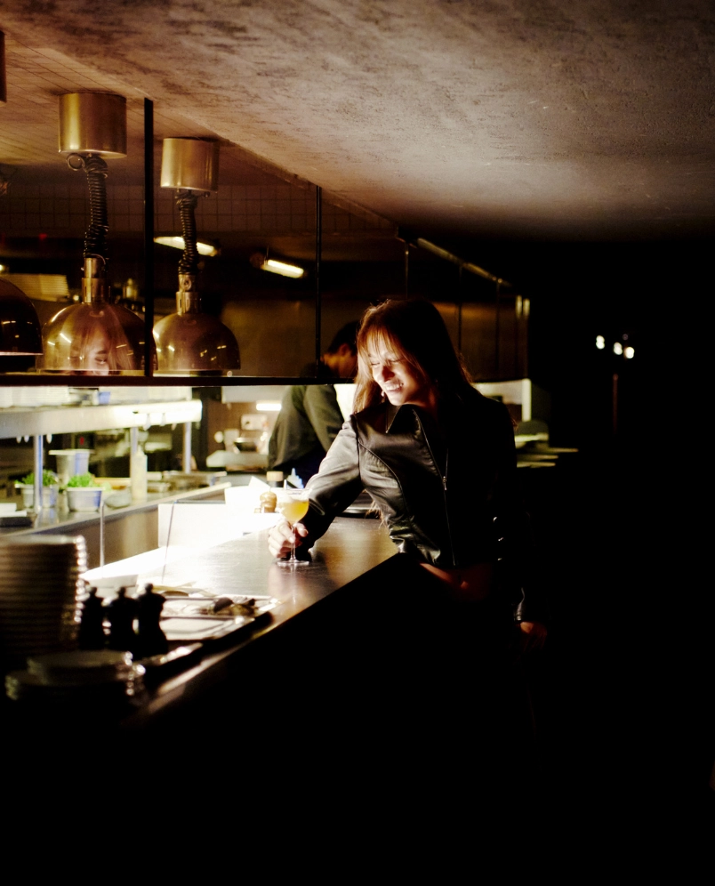 Smiling woman in a black jacket holding a cocktail glass at a dimly lit bar counter.