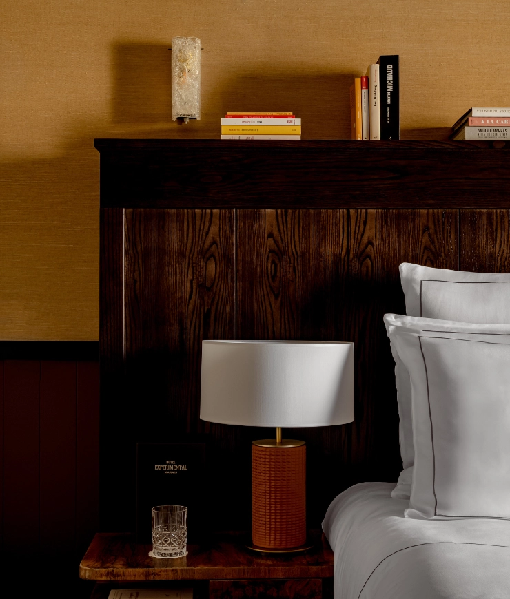 Bedside table with a textured orange lamp, crystal glass, and books against a dark wooden headboard with white pillows on the bed.