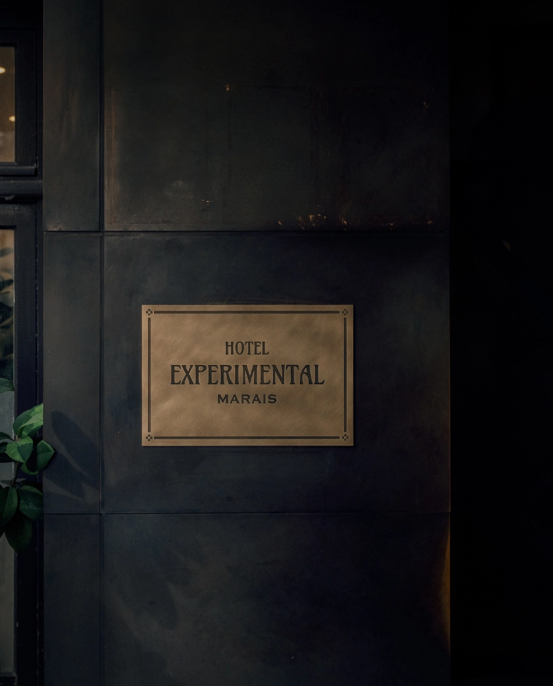Metal sign on dark wall reading 'Hotel Experimental Marais' with partial view of a plant on the left.