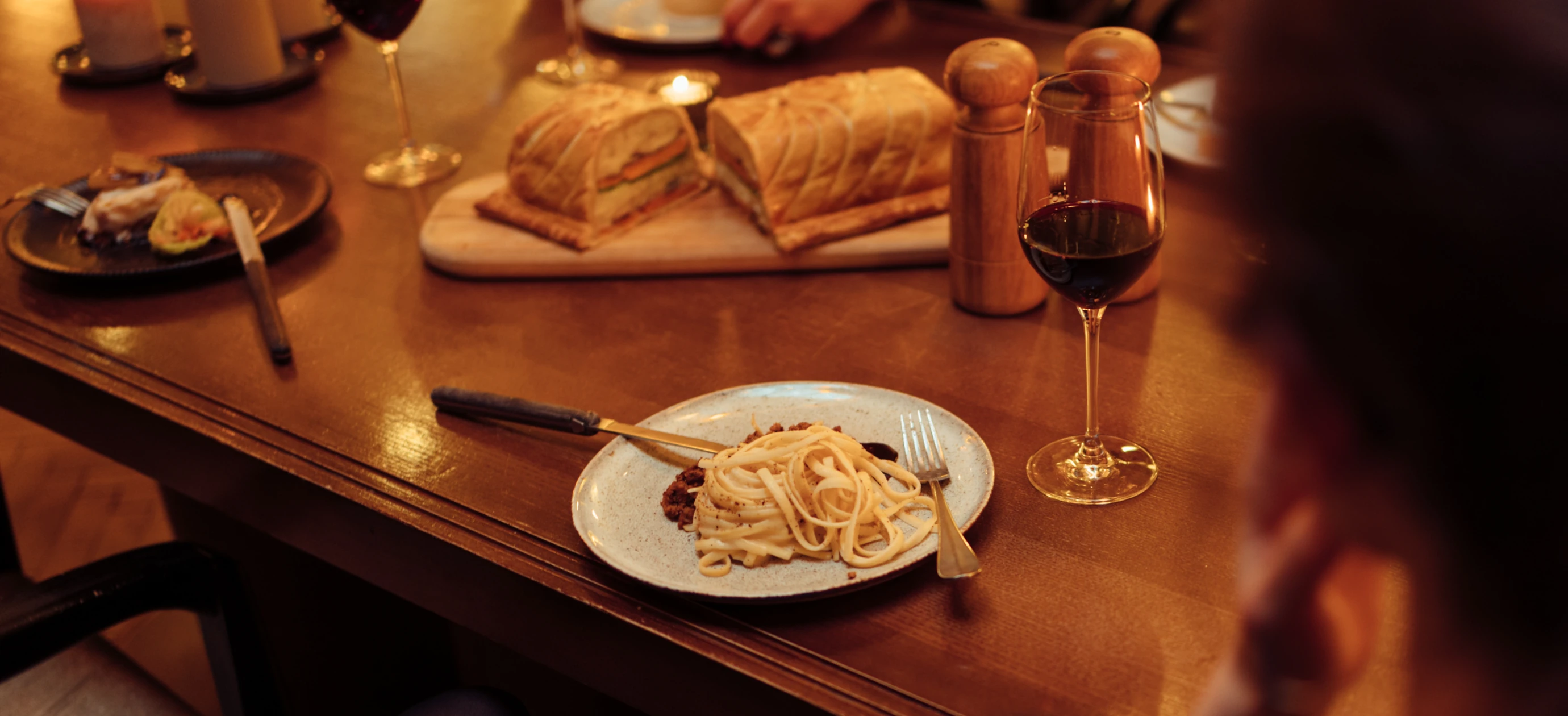 Plate of pasta with sauce on a wooden table next to a glass of red wine and bread loaves on a cutting board.