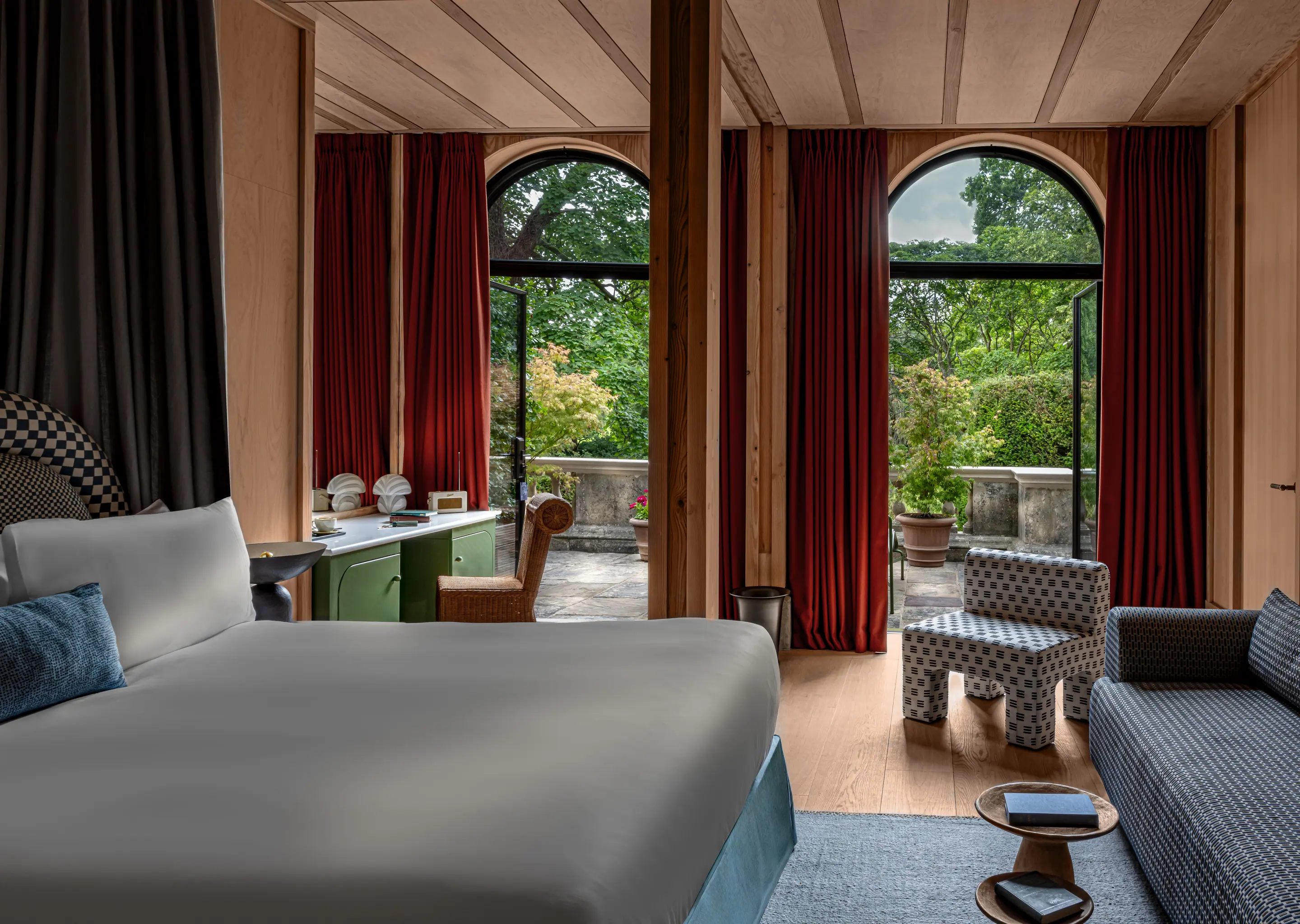 Modern hotel room with a large bed, patterned chair and sofa, wooden floors, and open arched doors leading to a green garden terrace.