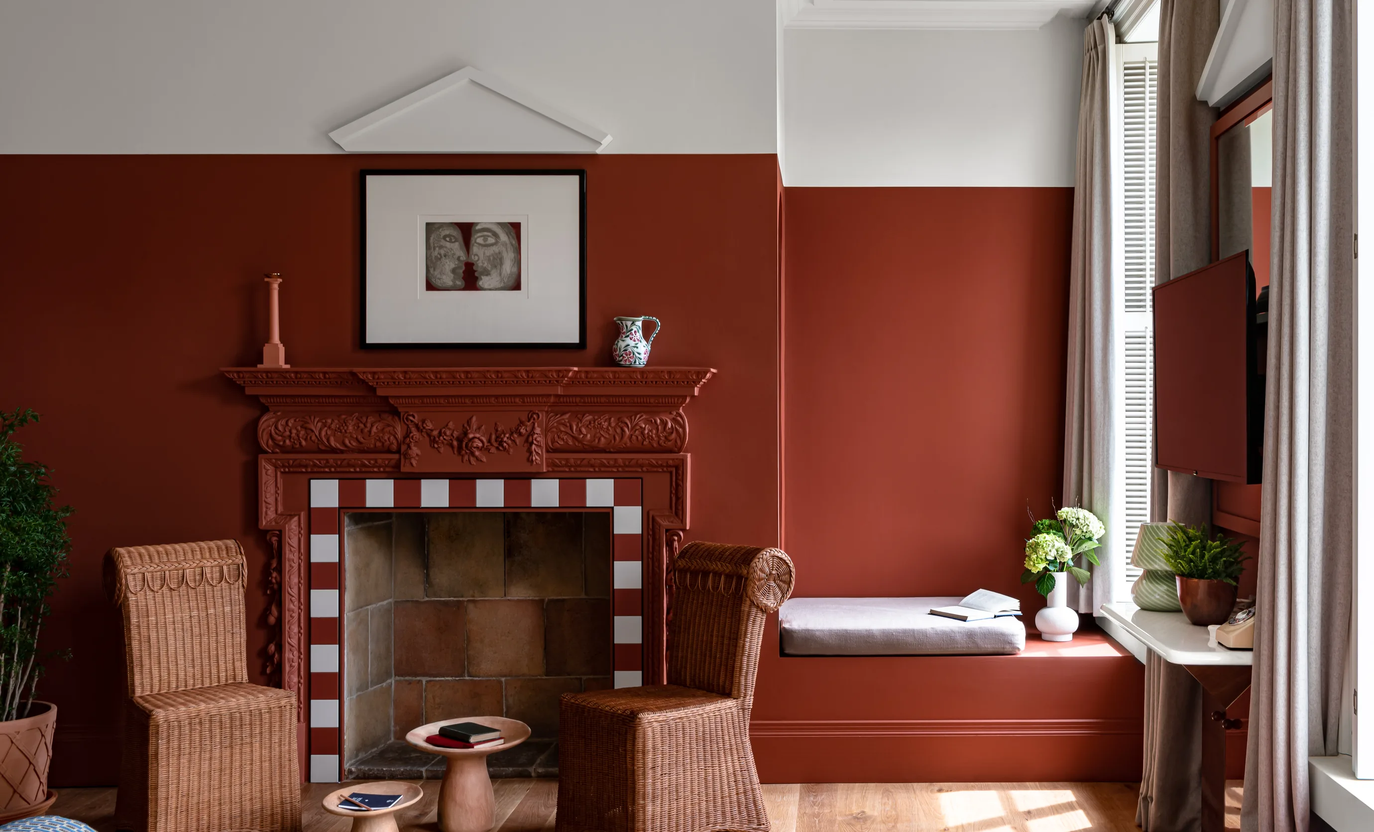 Cozy room with reddish-brown walls, ornate fireplace, two wicker chairs, a small wooden table, and a window seat with cushions and plants.