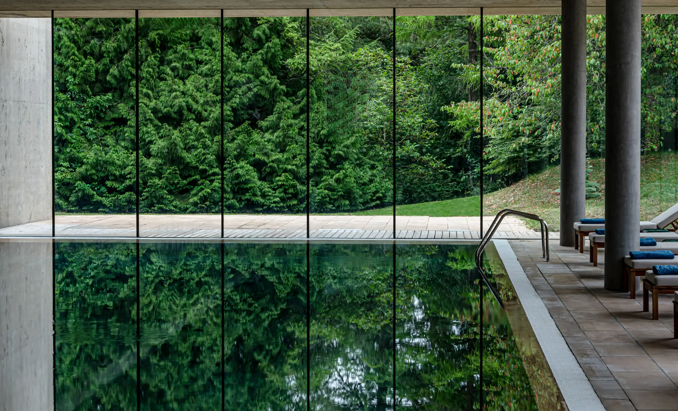 Indoor pool with glass wall reflecting lush green trees outside and lounge chairs with blue towels.