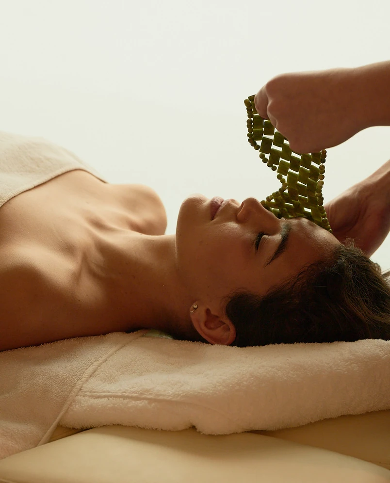Woman lying on a massage table with eyes closed while receiving a green bead facial massage.