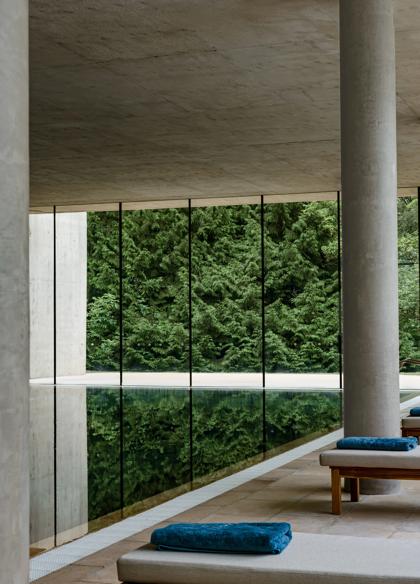 Indoor pool area with floor-to-ceiling windows showing green trees outside, concrete columns, and lounge chairs with blue towels.