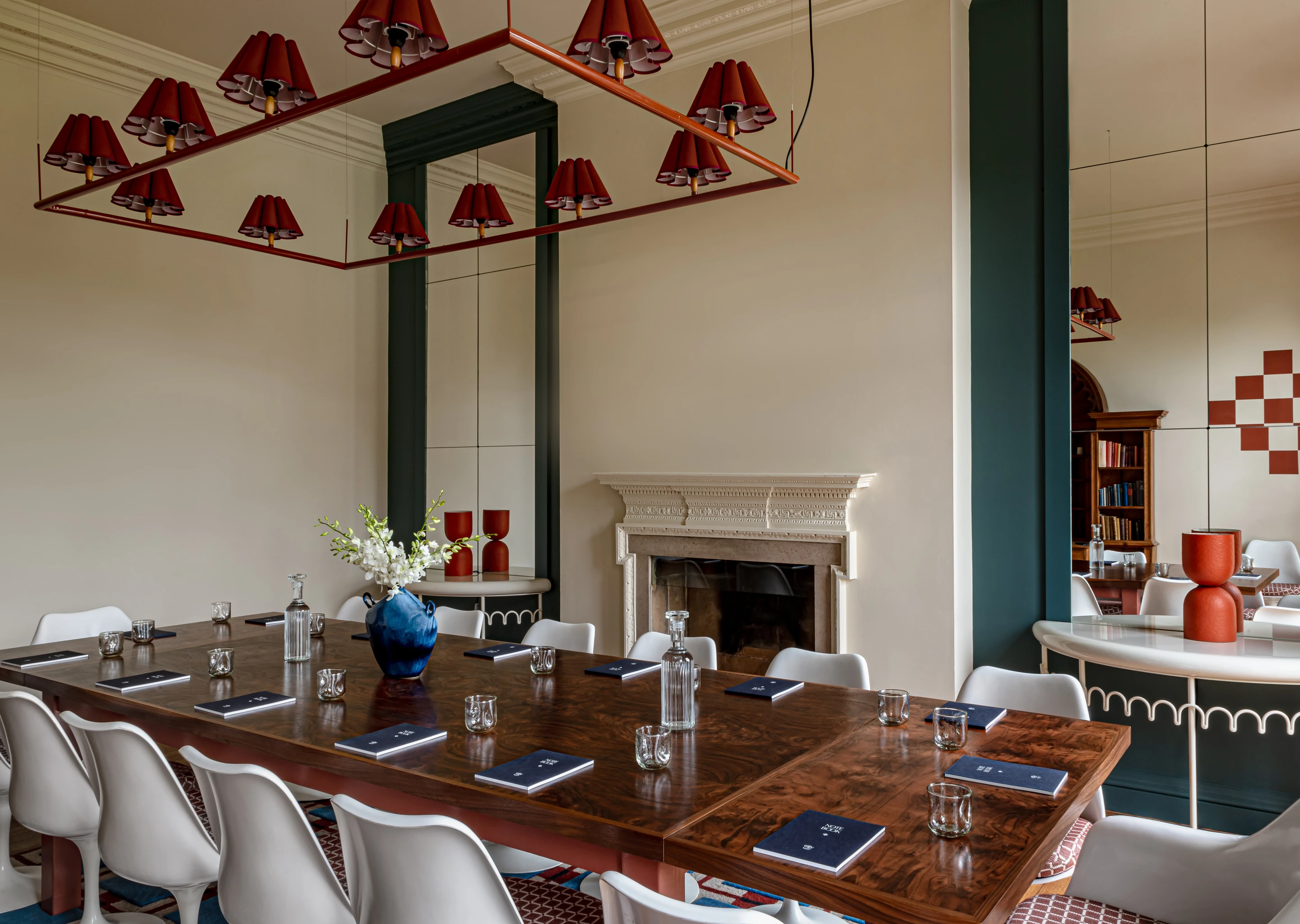 Meeting room with a large wooden table surrounded by white chairs, notebooks and glasses on the table, a blue vase with white flowers, and a decorative fireplace in the background.
