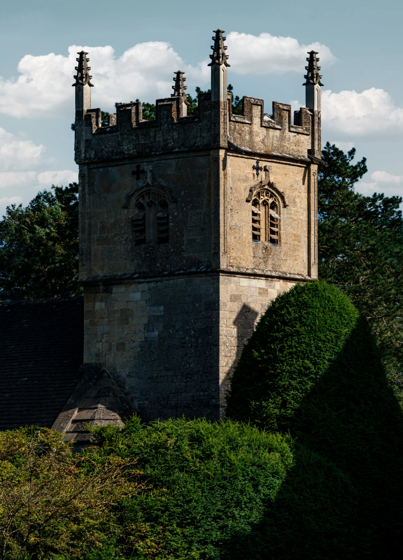 Stone tower with gothic windows and battlements surrounded by green shrubs and trees under a partly cloudy sky.