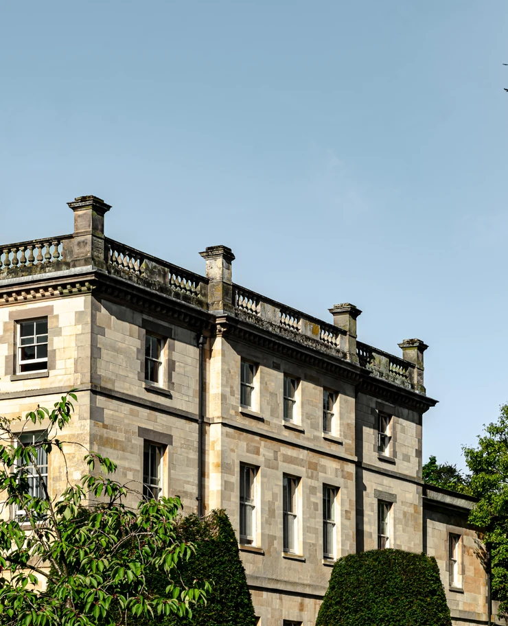 Stone building with classical columns and multiple windows under a clear blue sky, with green trees and bushes in front.