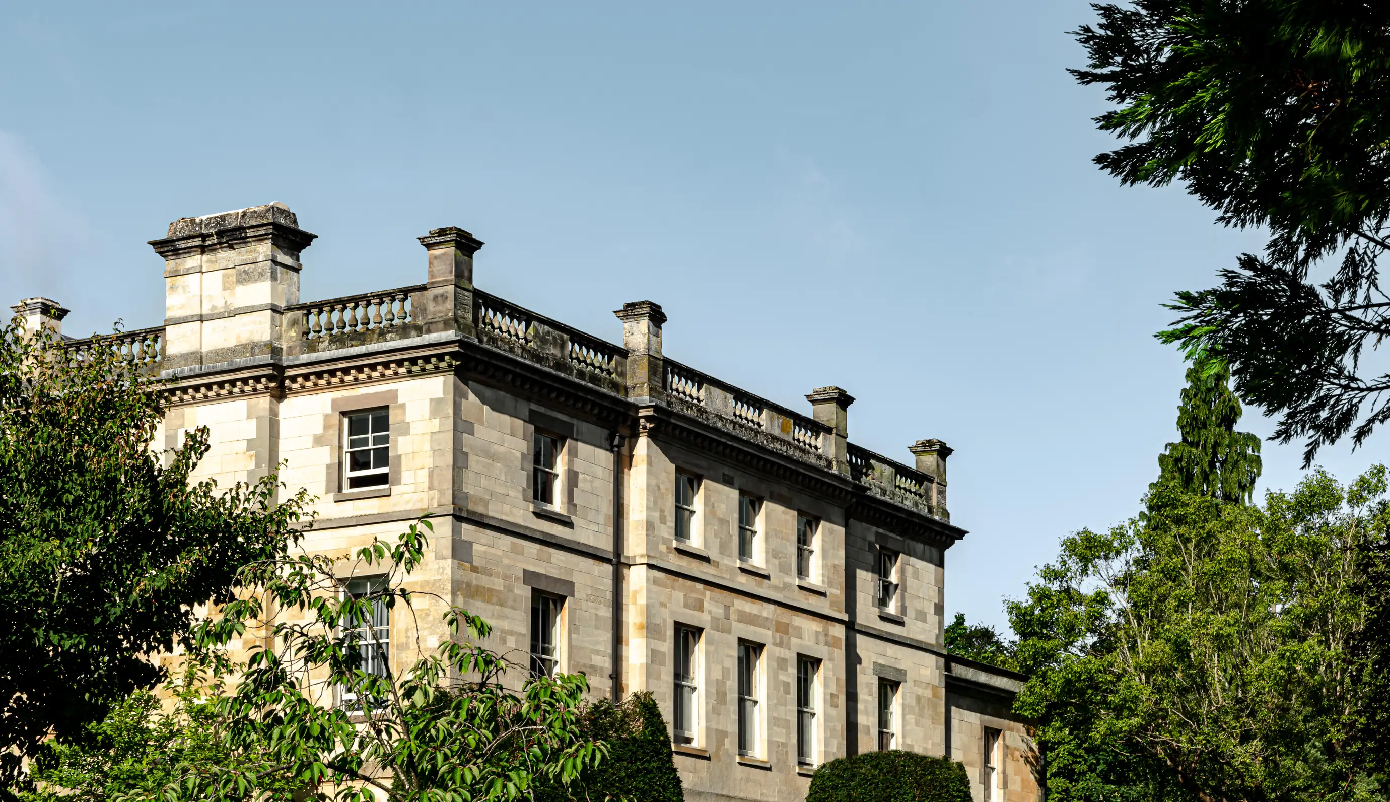 Stone building with classical architectural details surrounded by green trees and bushes under a clear sky.
