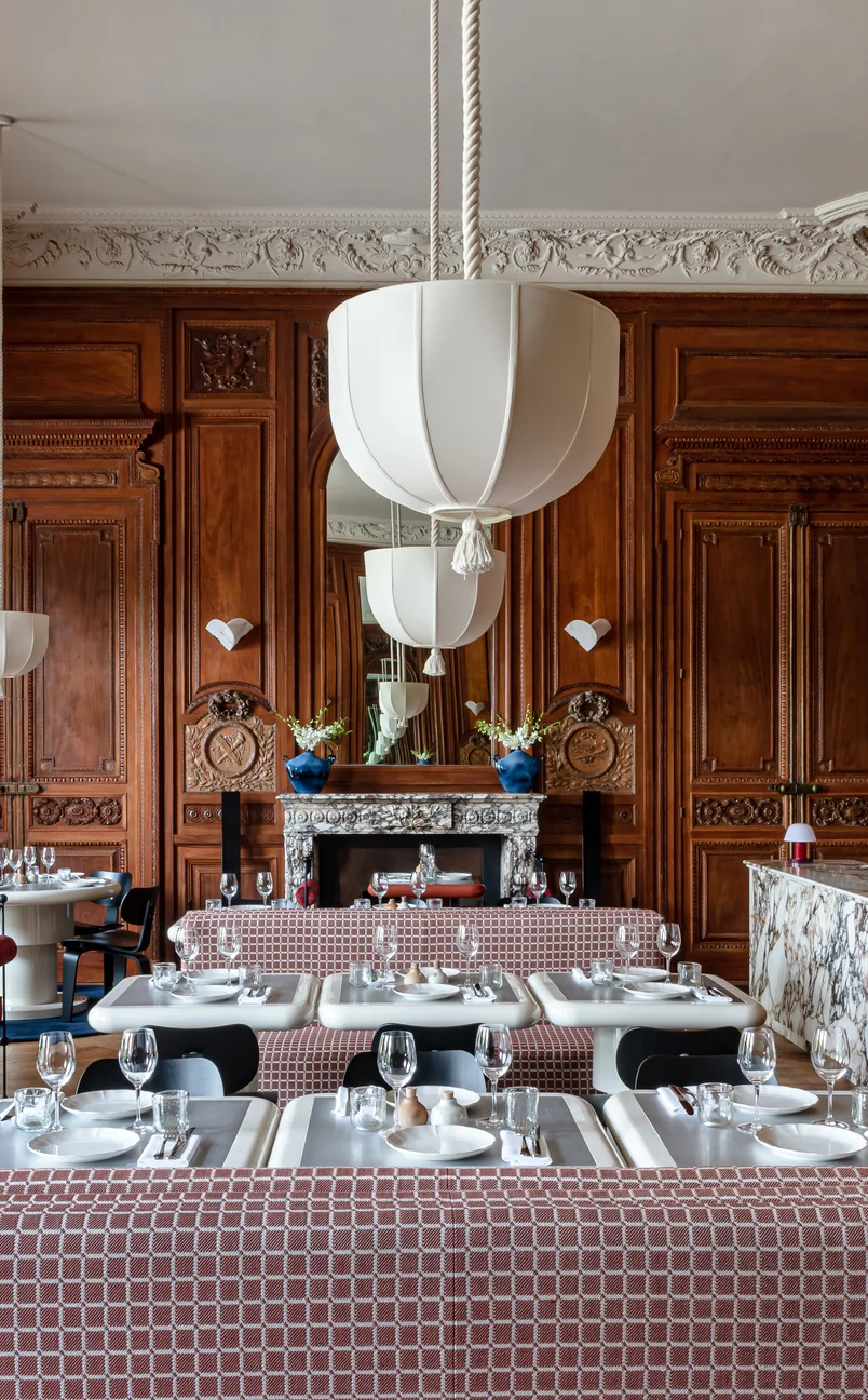 Elegant restaurant interior with wooden paneled walls, white hanging lamps, marble fireplace, and neatly set tables with patterned bench seating.