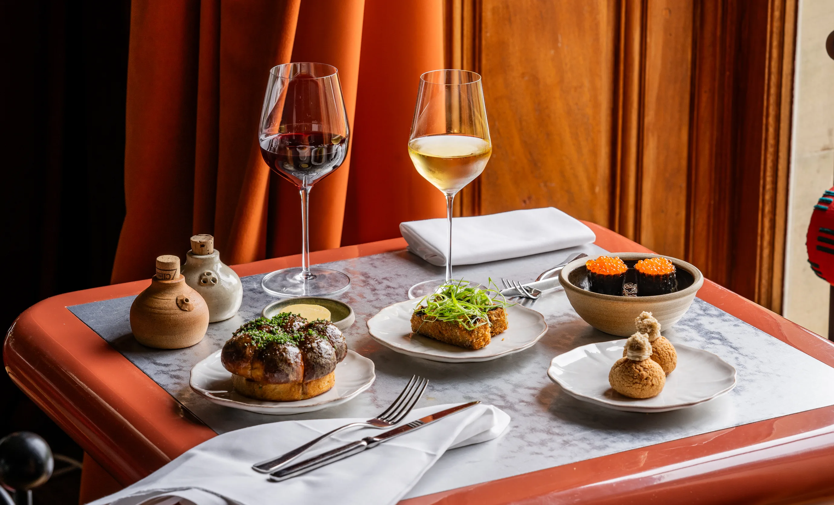 Table set with gourmet dishes including bread roll, fried square topped with greens, sushi with orange roe, two cream-filled pastries, and glasses of red and white wine.