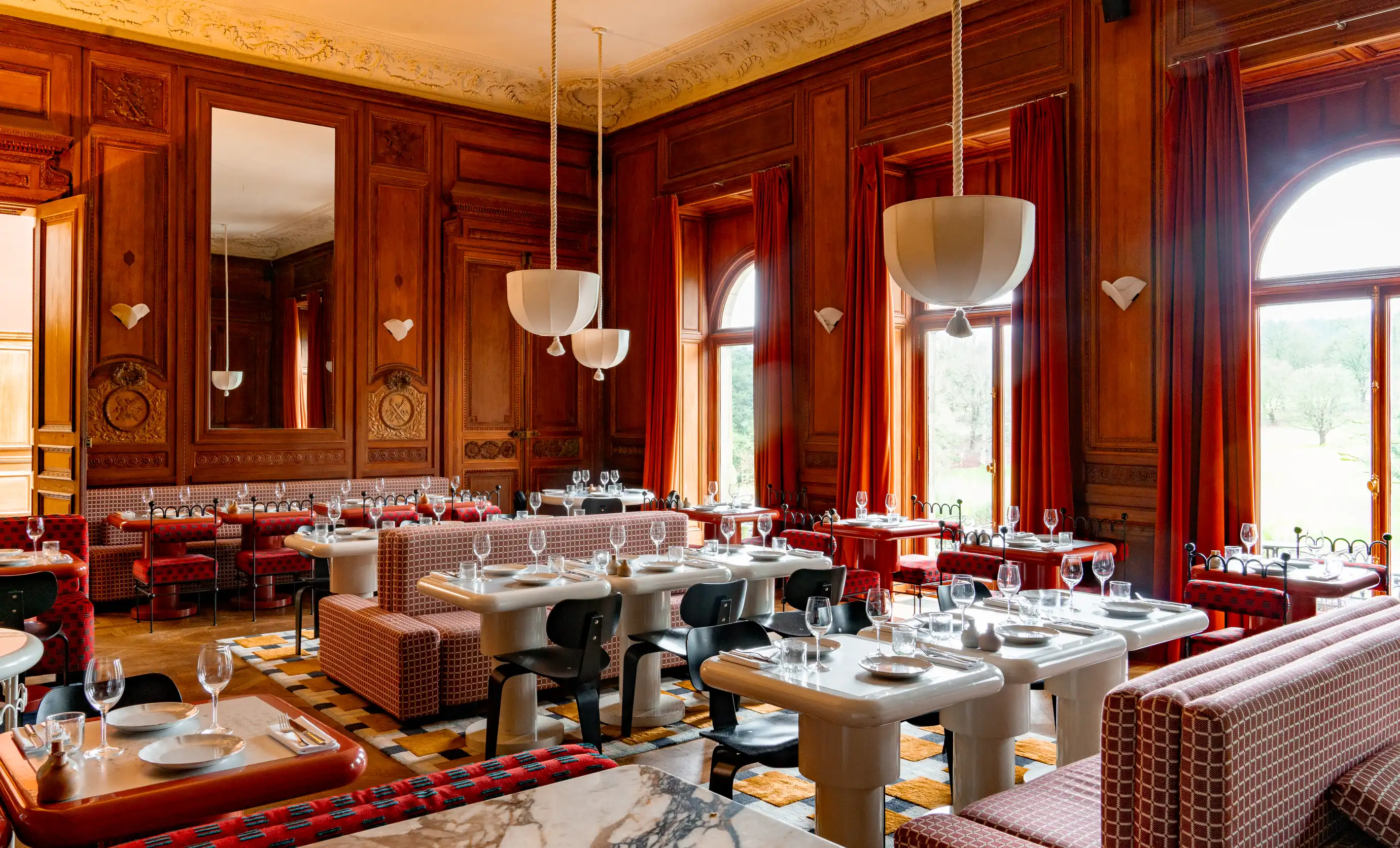Elegant restaurant interior with wooden paneling, large arched windows with red curtains, and neatly set tables with white plates and glassware.