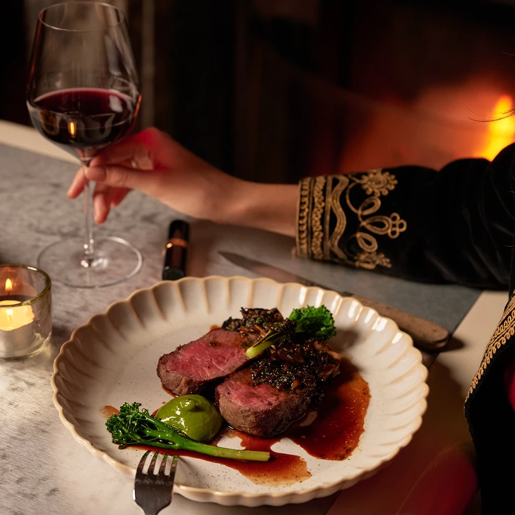 Plate with two slices of medium-rare steak, green vegetables, and sauce on a marble table next to a hand holding a glass of red wine and a lit candle.