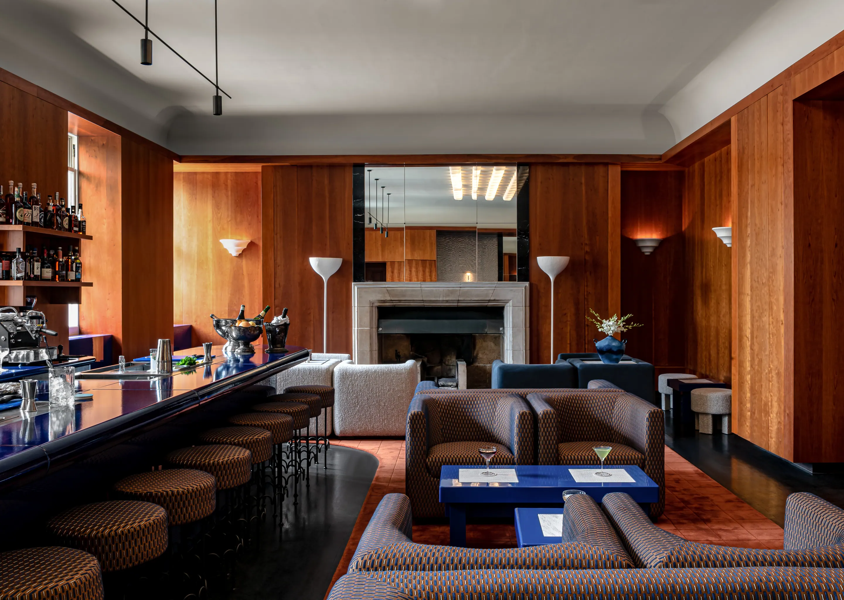 Modern cocktail bar with a long navy blue counter, patterned stools, plush armchairs, and a fireplace framed by wood-paneled walls.