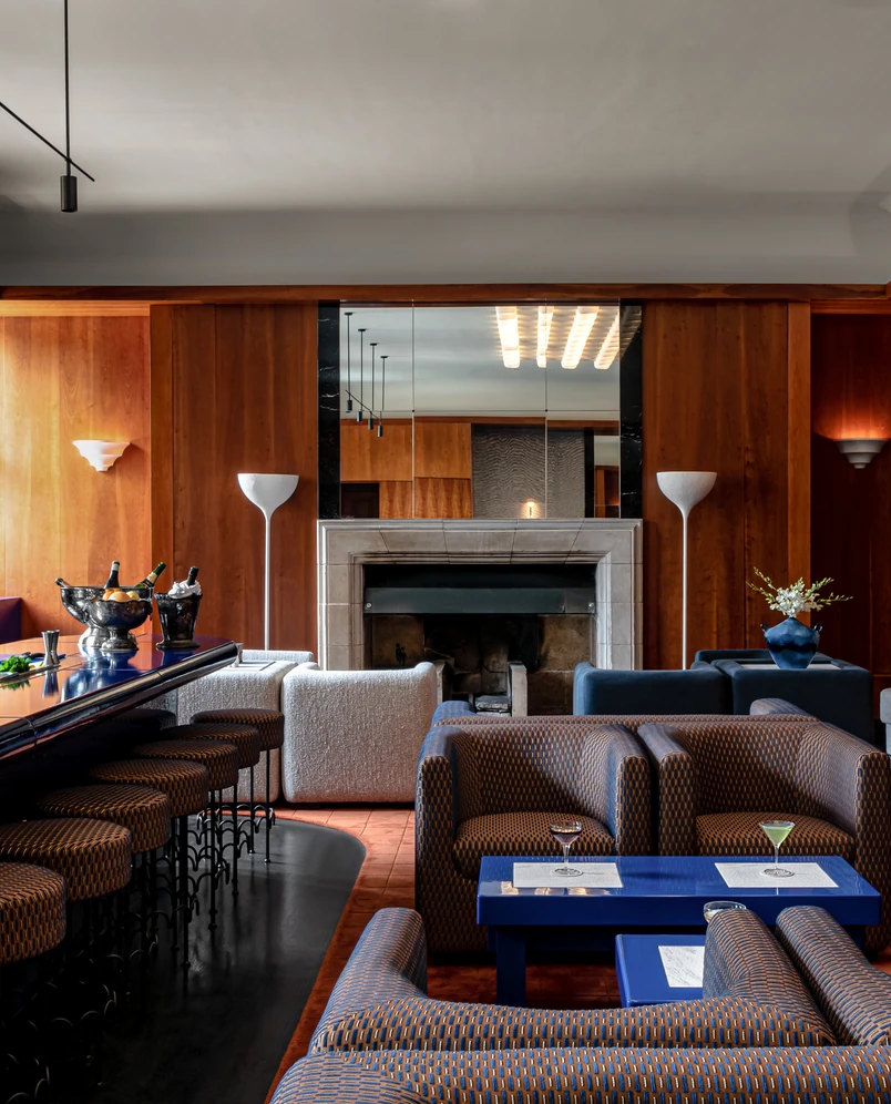 Modern cocktail bar interior with patterned armchairs, blue tables, a marble fireplace, wood-paneled walls, and a bar with stools and champagne buckets.
