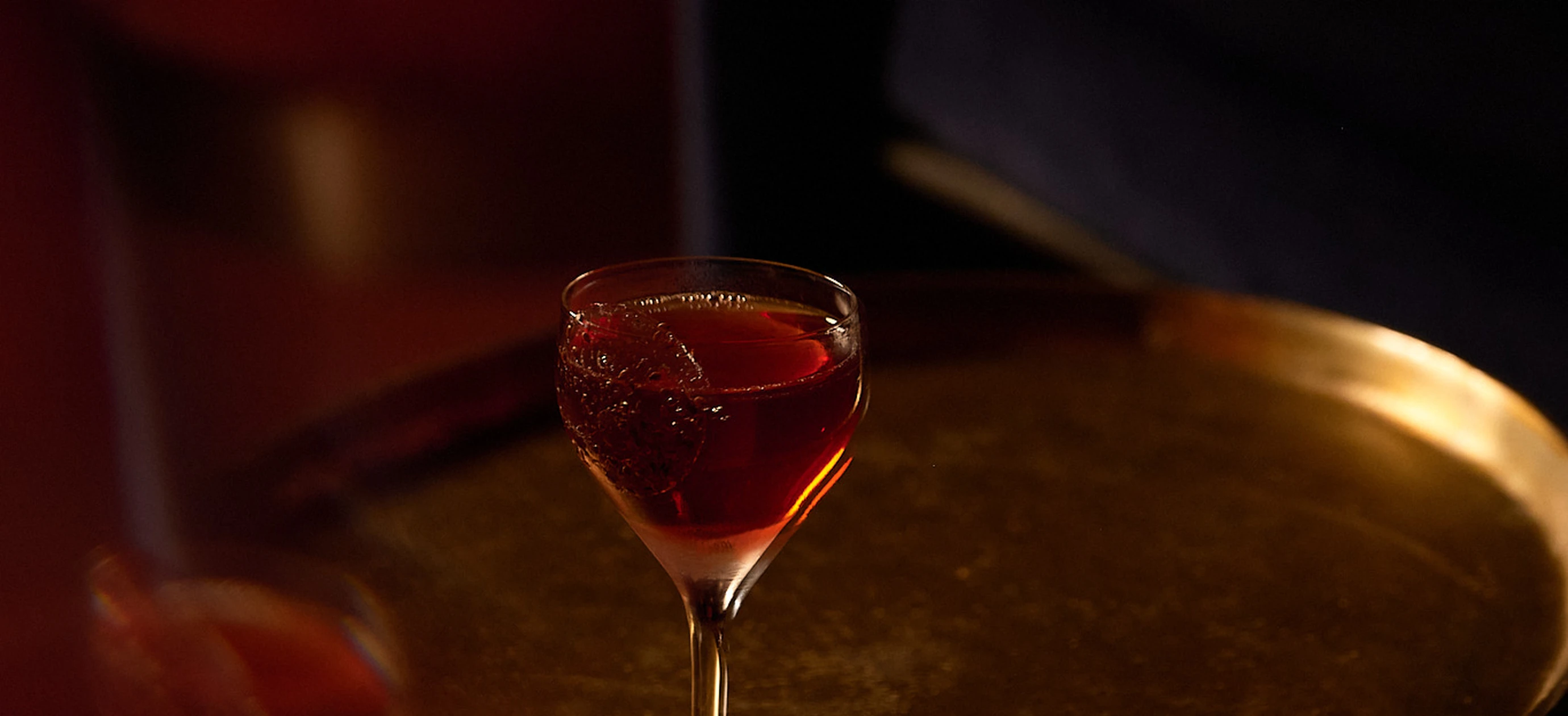 A glass of dark reddish cocktail on a round brass tray with dim warm lighting.