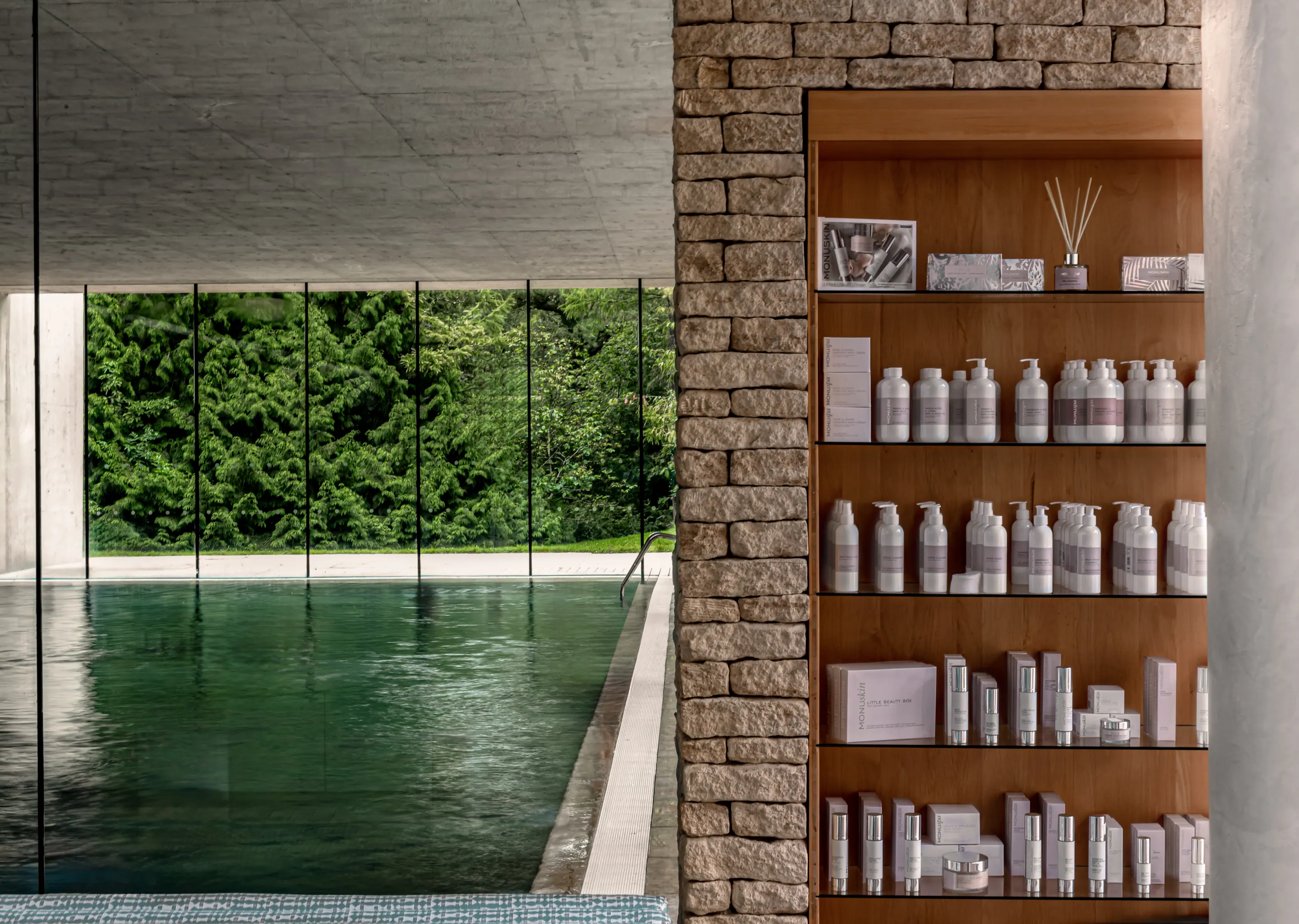 Indoor pool area with large windows showing green trees outside and a wooden shelf filled with skincare products.