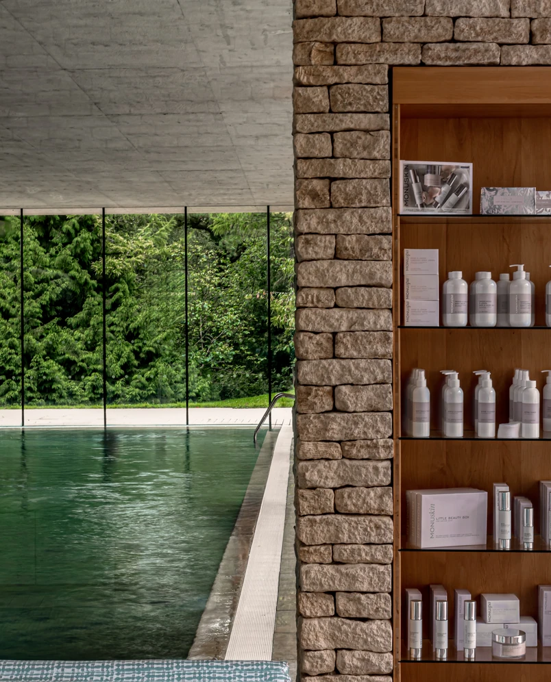 Indoor pool area with a stone wall and wooden shelves displaying skincare and beauty products, large windows show lush green trees outside.