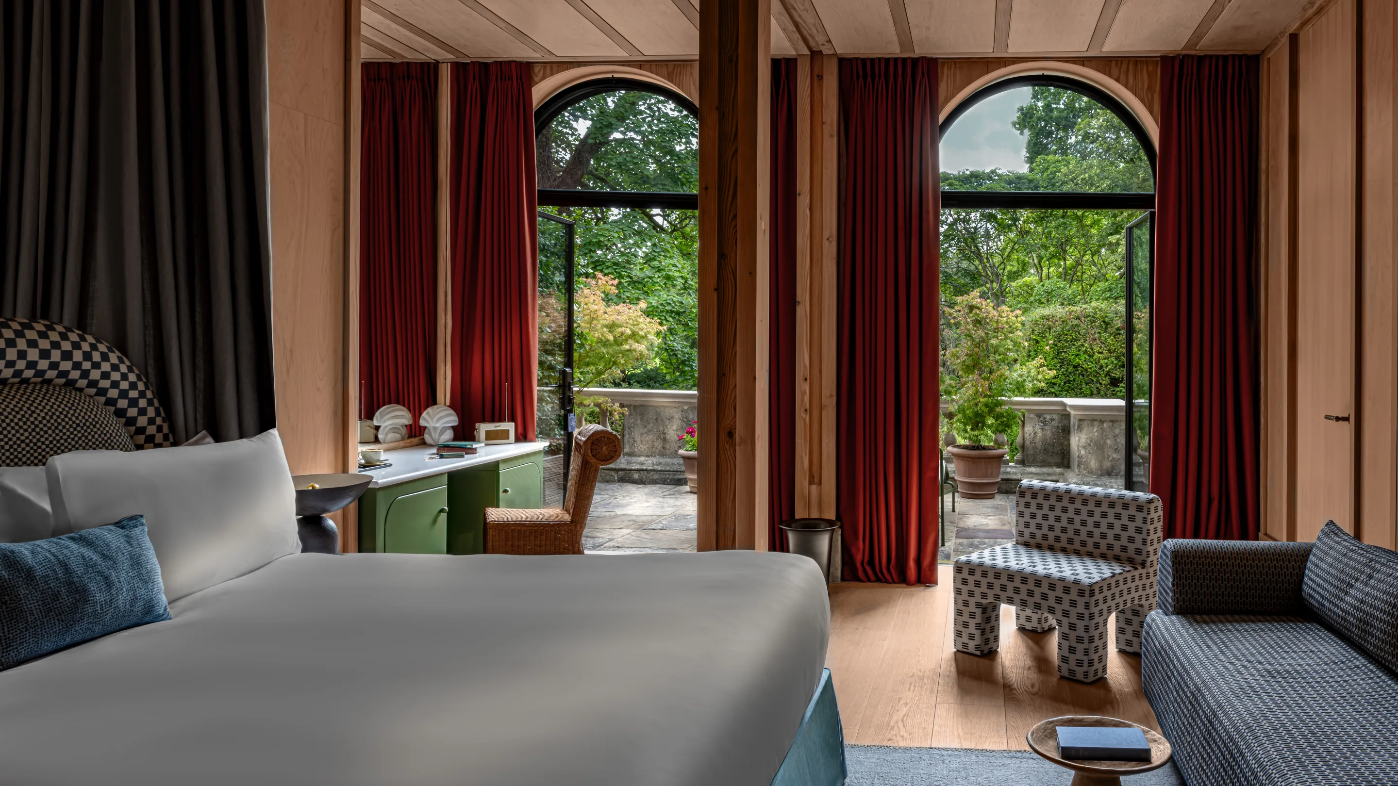 Hotel room with a large bed, patterned chairs, red curtains, and open arched doors leading to a green garden terrace.