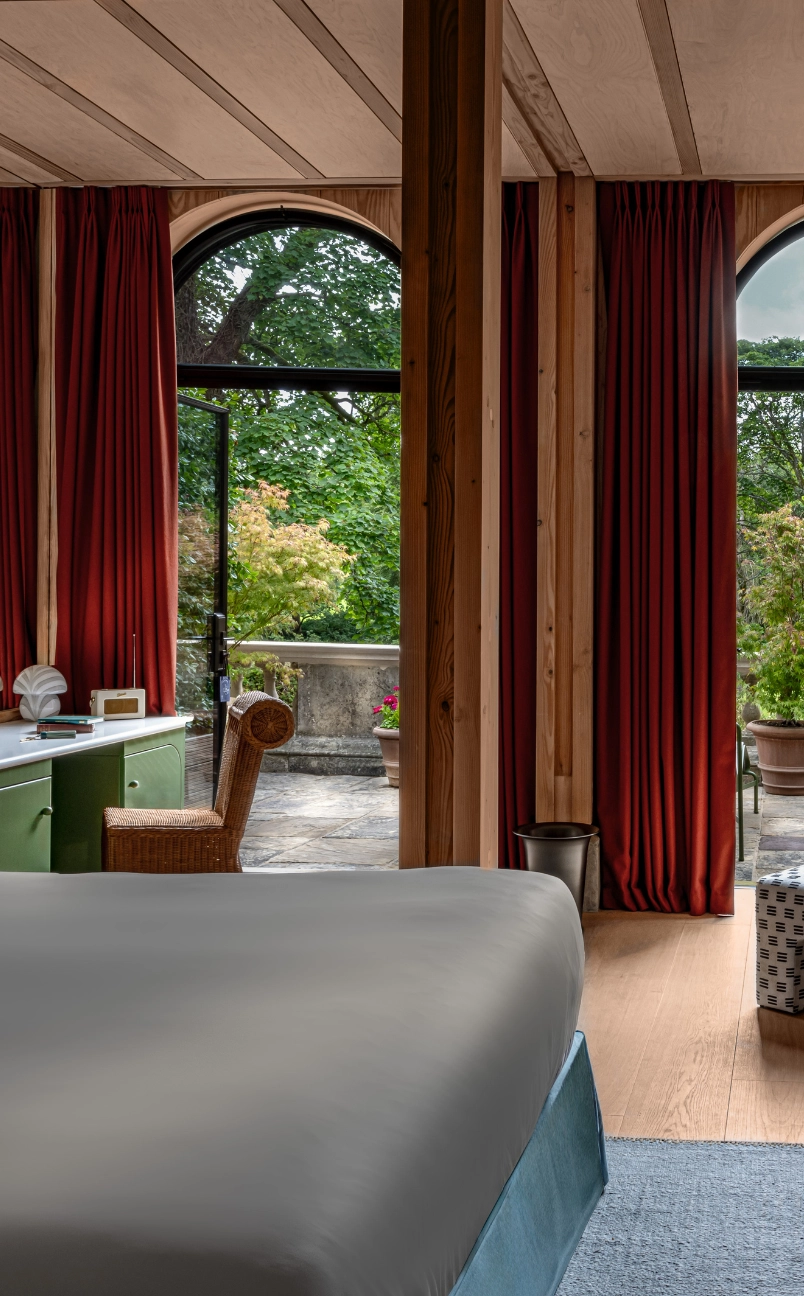 Hotel room with a large bed, green desk, wicker chair, and arched windows with red curtains overlooking a leafy outdoor terrace.