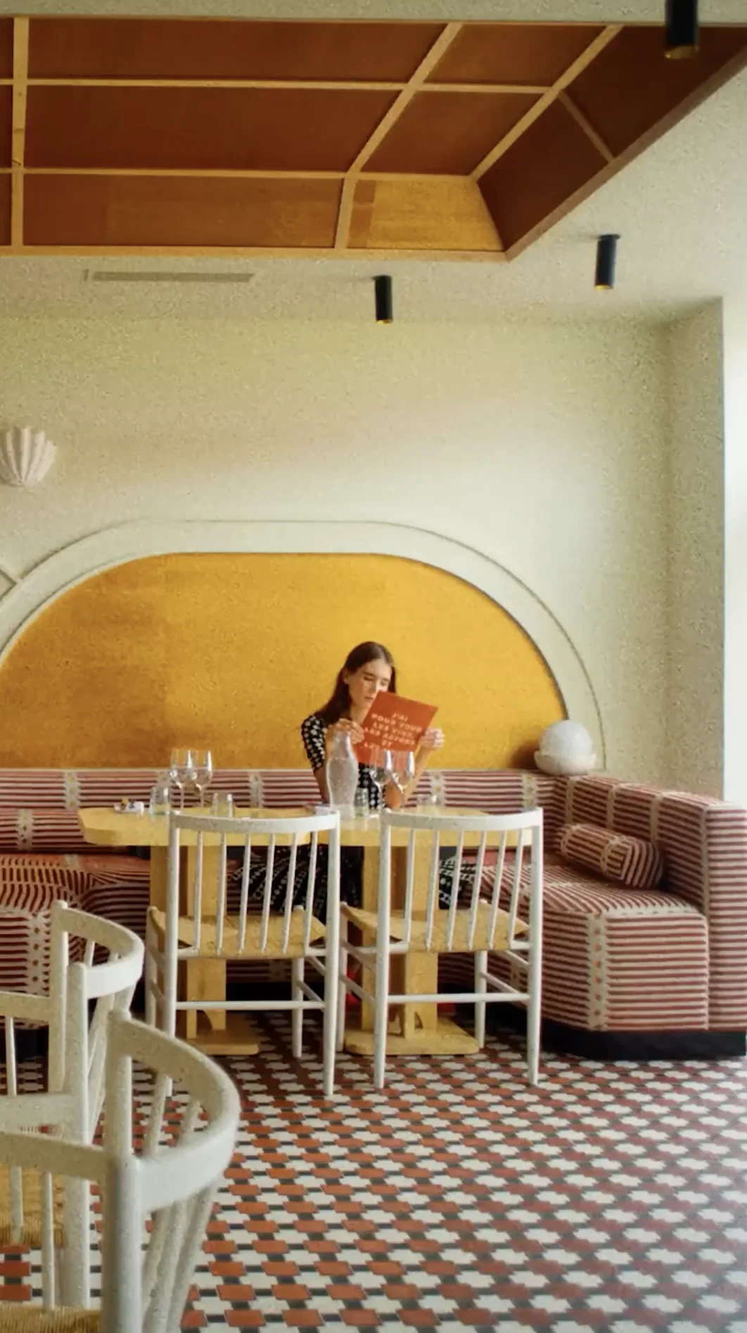 Woman sitting at a table in a patterned floor restaurant reading a menu, with striped cushioned benches and wooden chairs around.