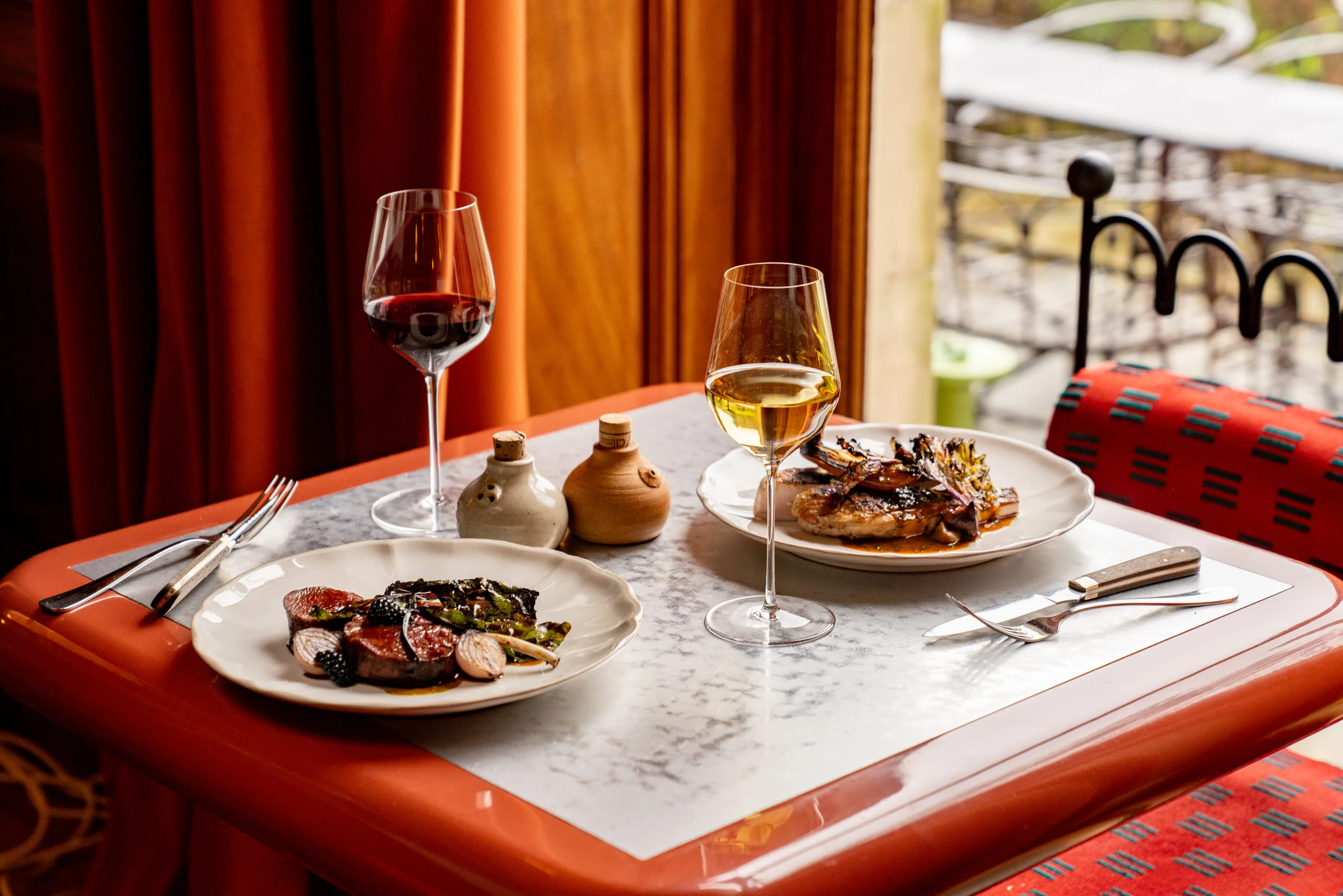Table set for two with plates of gourmet food and glasses of red and white wine in a cozy restaurant setting.