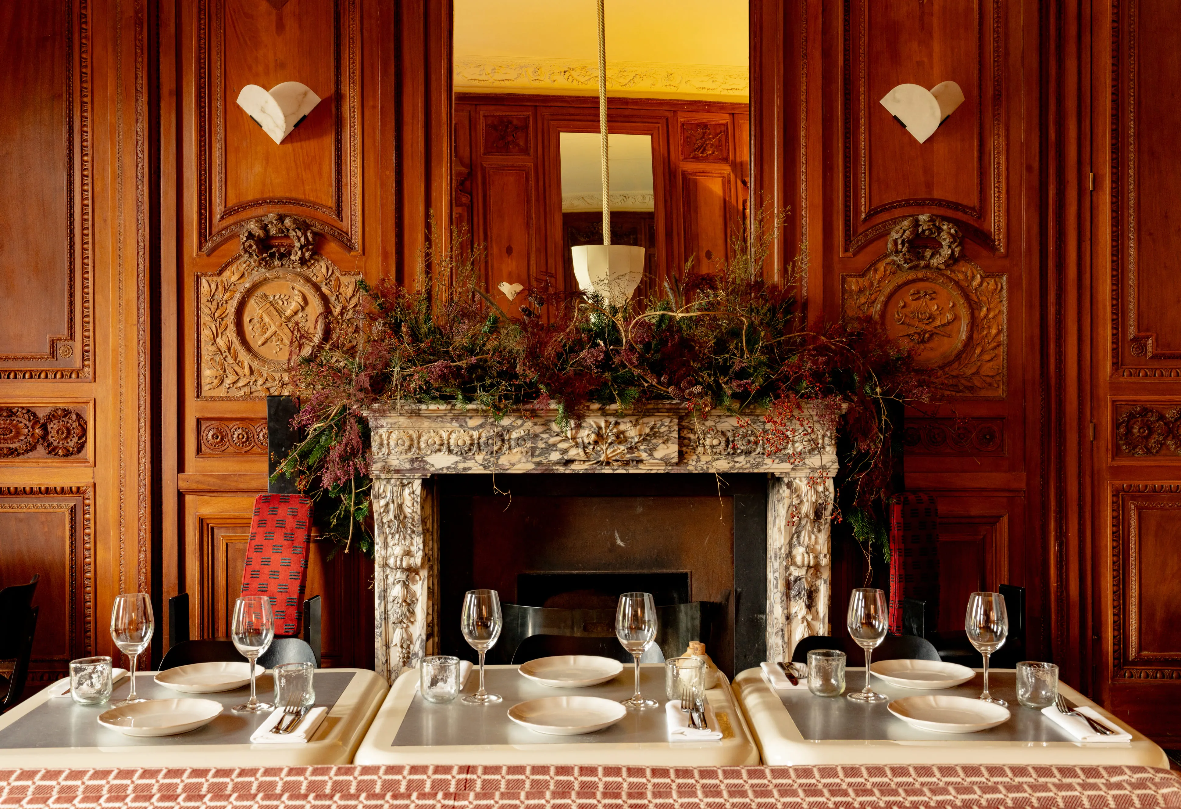Elegant dining setup with white plates, wine glasses, and silverware on tables in front of an ornate marble fireplace decorated with lush foliage and surrounded by carved wooden paneling.