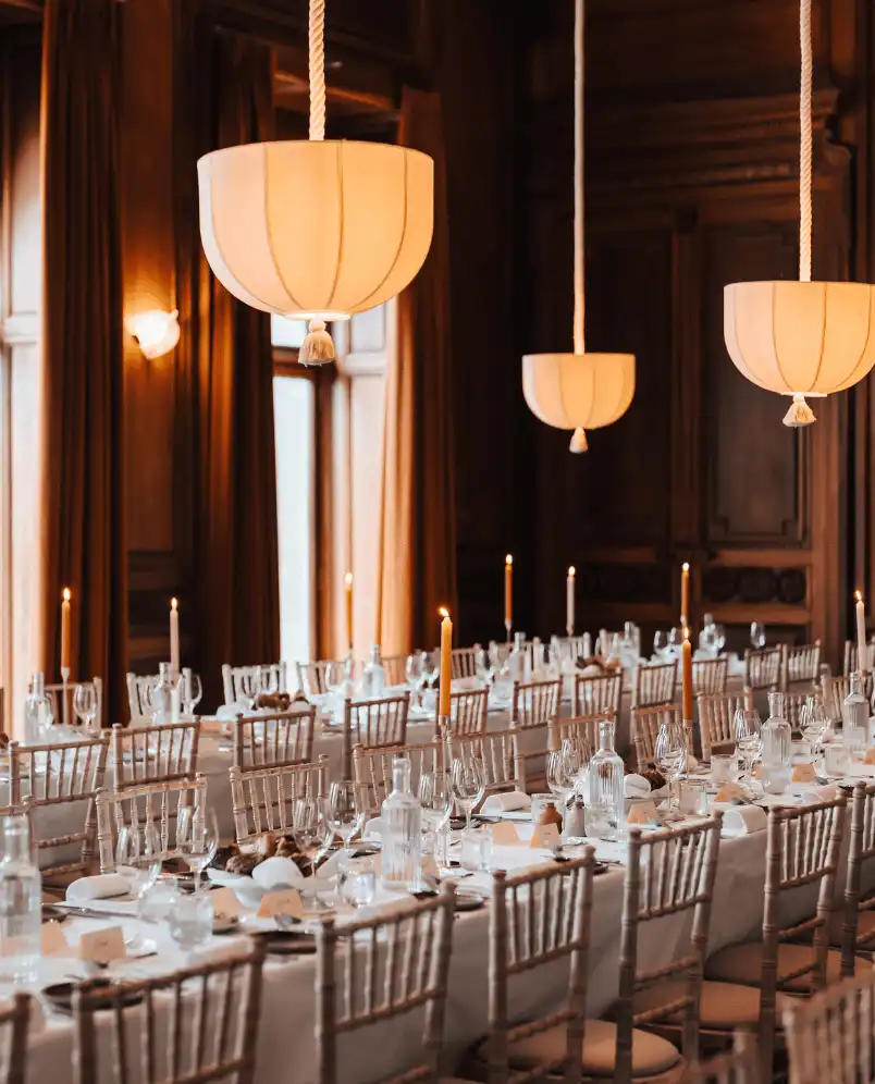 Long dining tables set for an elegant event with white tablecloths, glassware, candles, and wooden chairs in a warmly lit room.