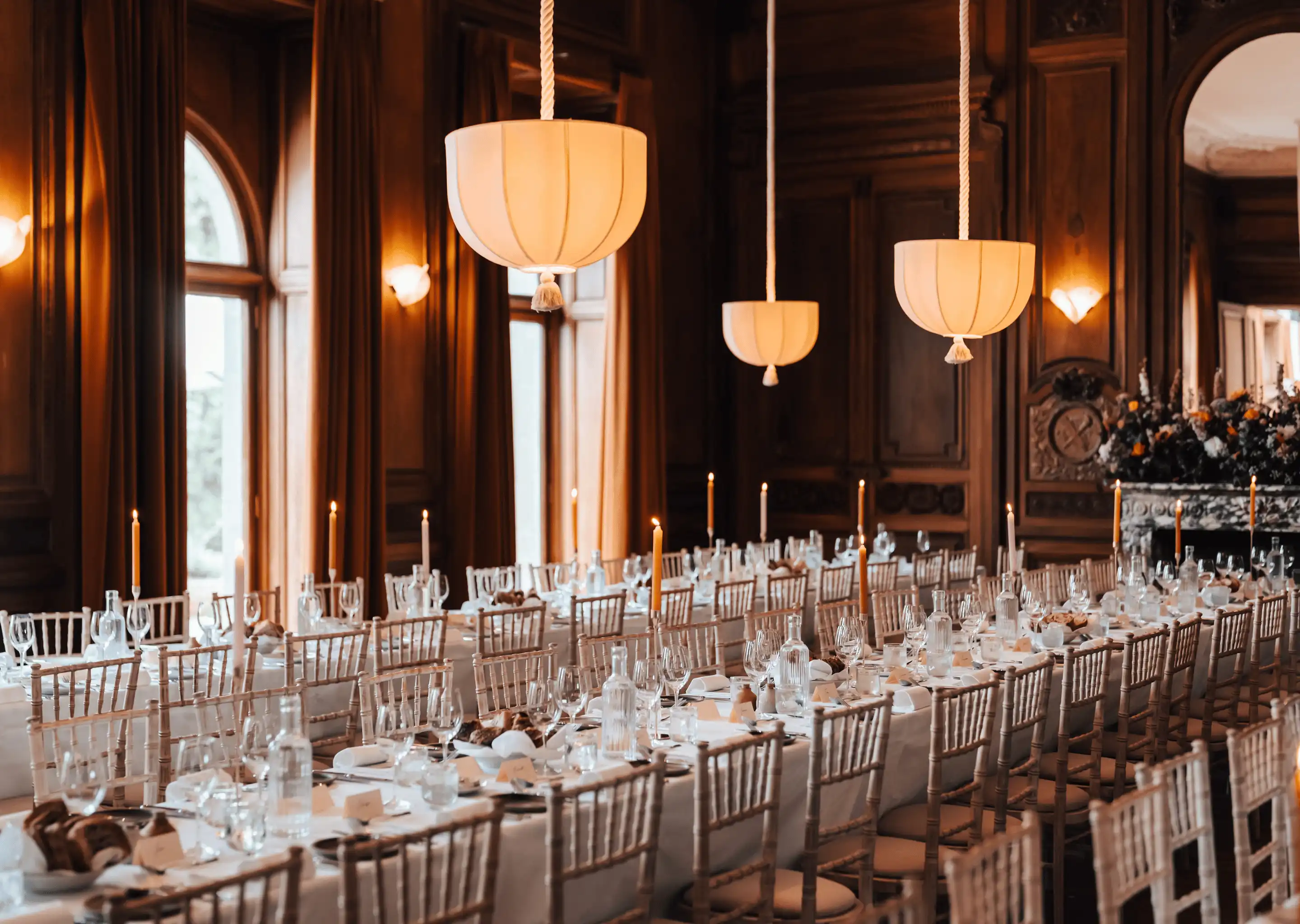Long dining tables set for a formal event with candles, glassware, and wooden chairs in an elegant wood-paneled room.