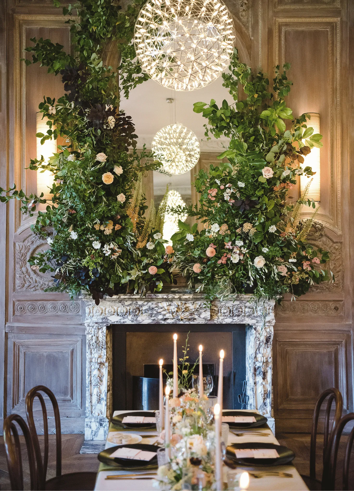 Elegant dining table with candles set in front of a marble fireplace adorned with lush greenery and flowers under a glowing spherical chandelier.