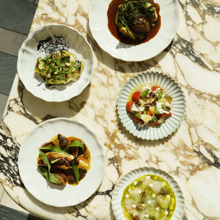Five plates of gourmet dishes including greens, roasted vegetables, and sauces arranged on a marble table.