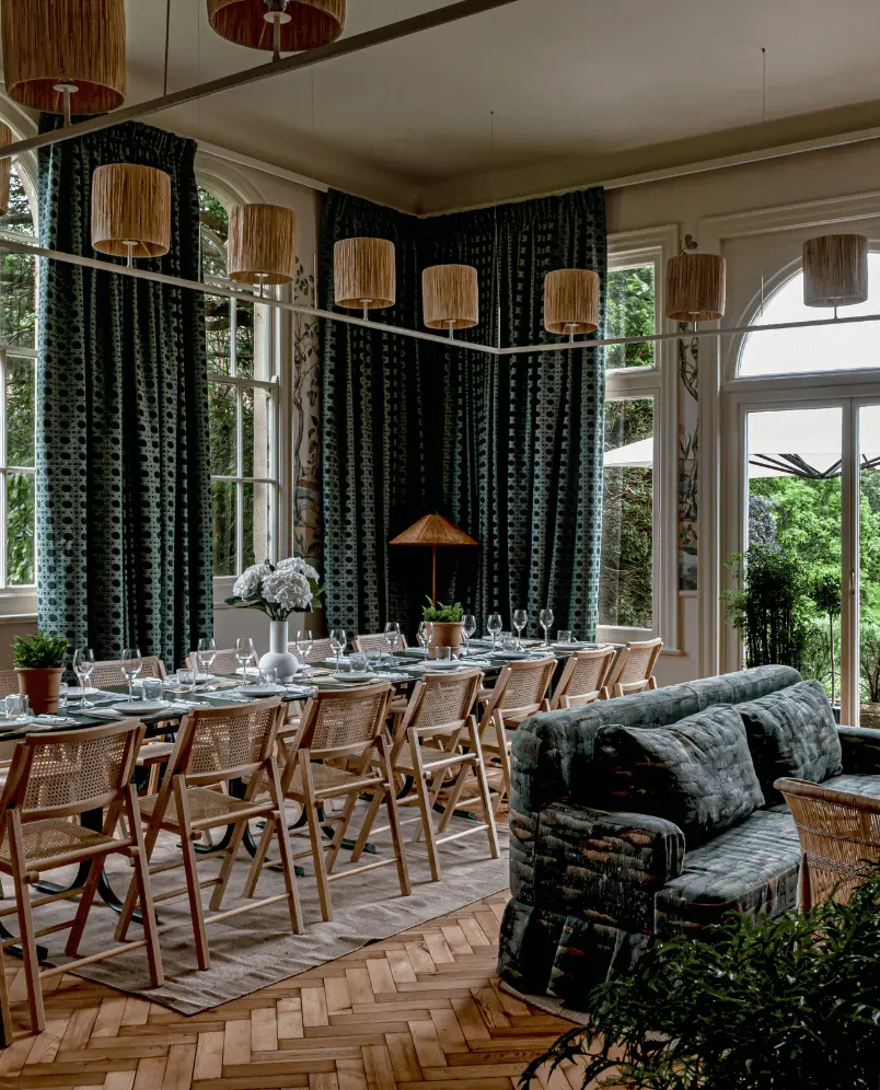 Elegant dining area with a long wooden table set with glasses and plates, surrounded by wooden chairs and a dark patterned sofa, under hanging wicker lamps.