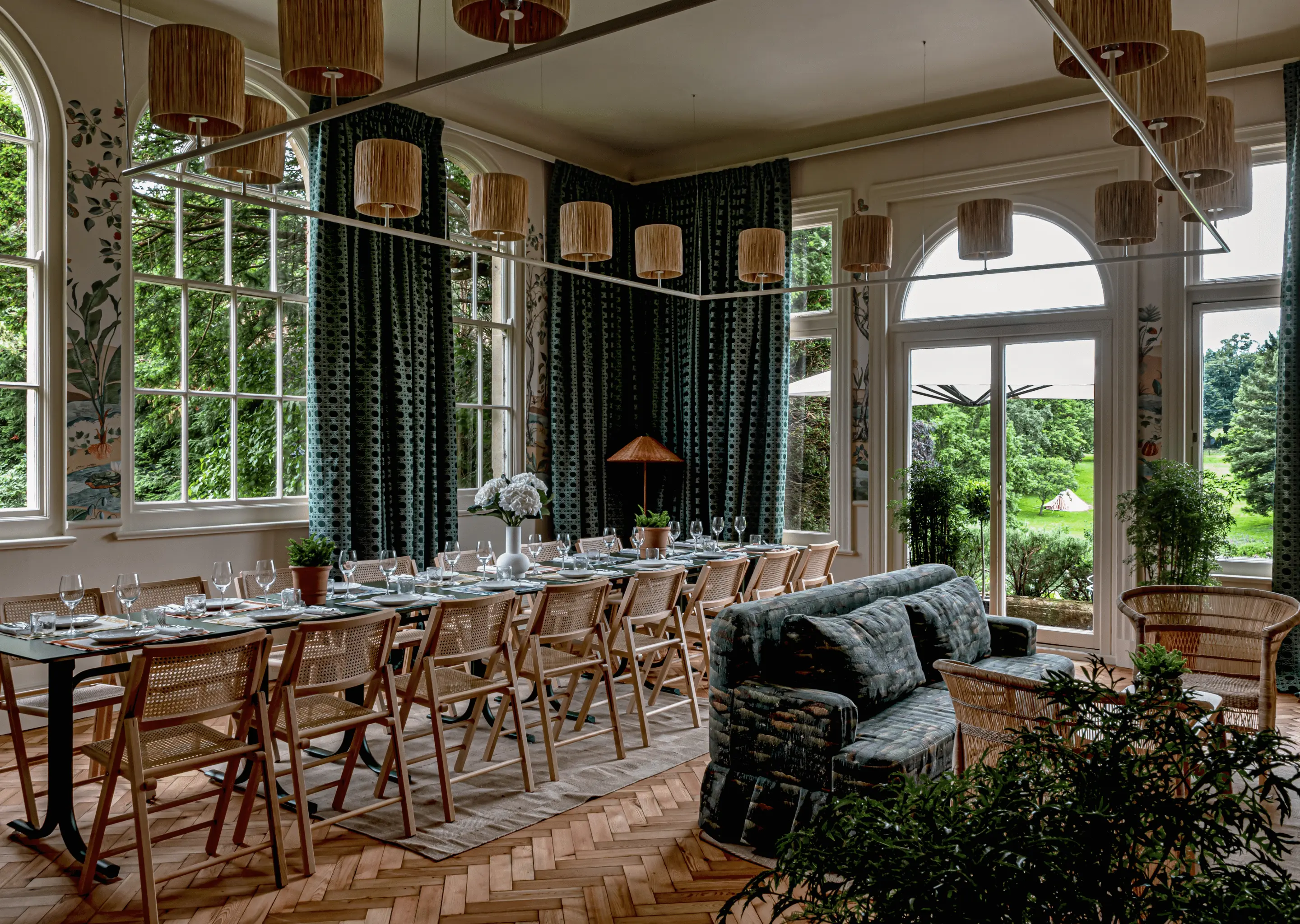 Elegant dining room with long table set for a meal, wicker chairs, patterned curtains, and large windows overlooking a garden.