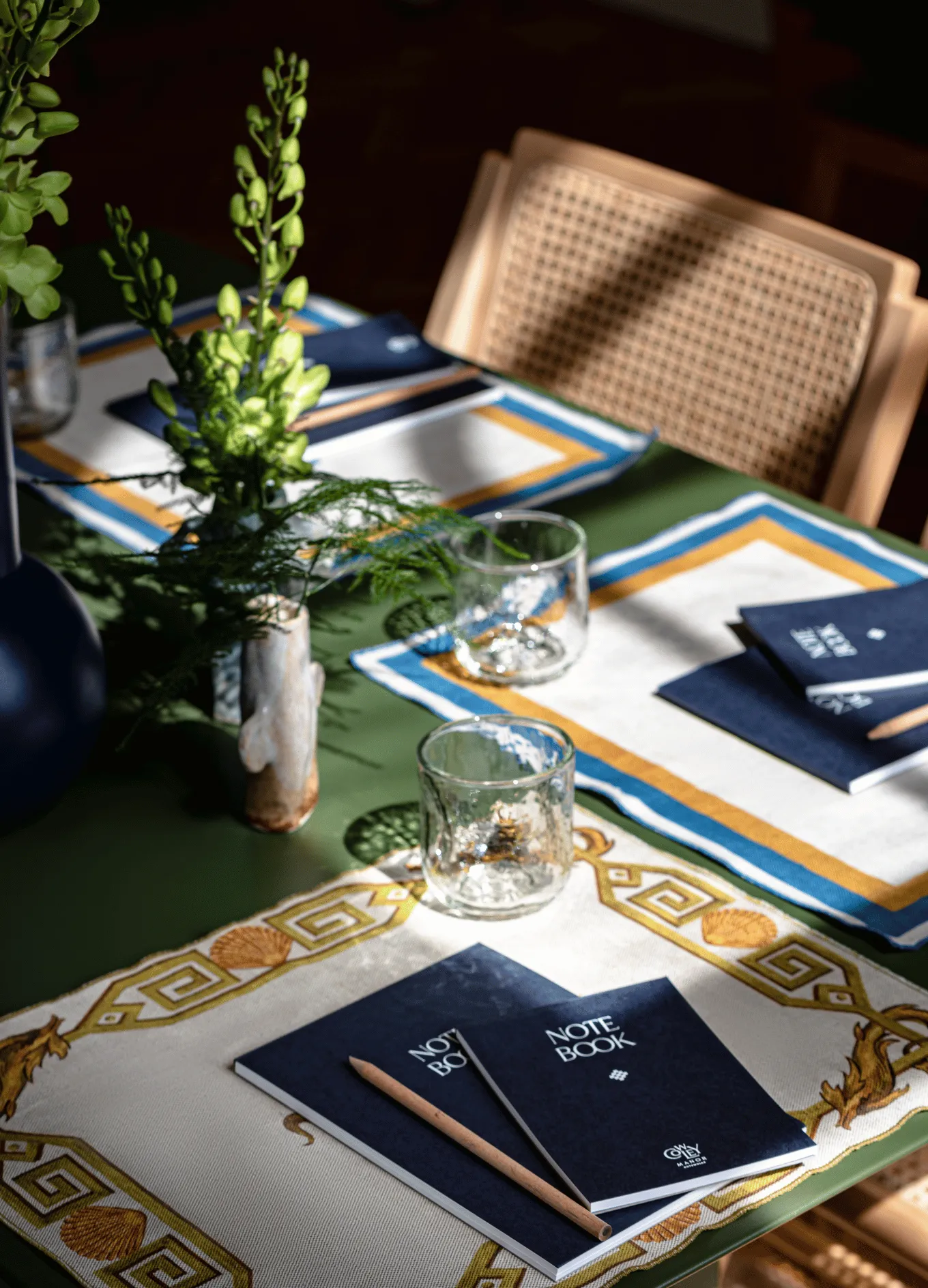 Table set with decorative placemats, two notebooks with pencils, clear glasses, and a small vase of green plants beside a woven chair.