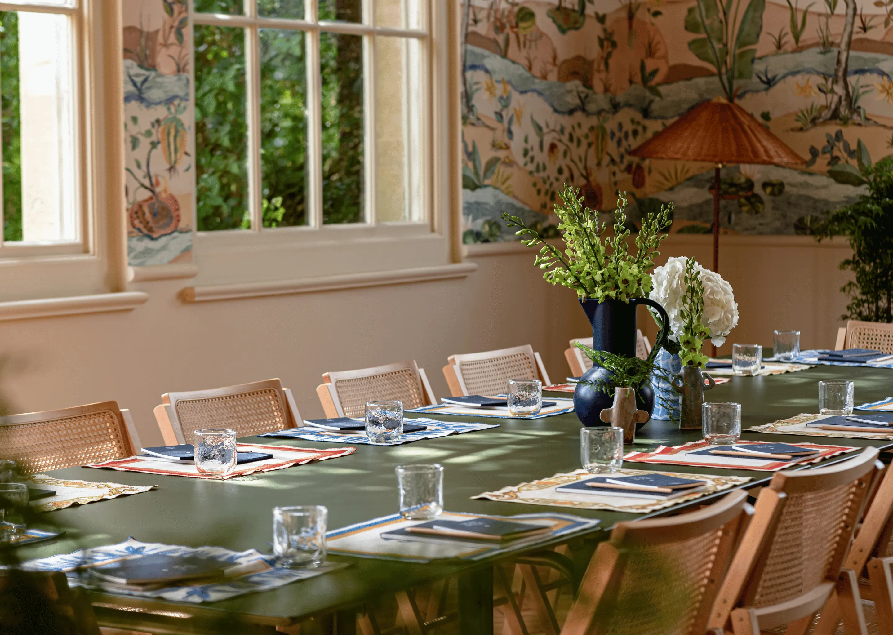 Long green table set with placemats, notebooks, and glasses, surrounded by wooden chairs in a room with large windows and botanical wallpaper.
